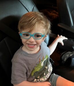 Toddler boy wearing a grey shirt and blue glasses smiling while sitting in a car with black leather seats.
