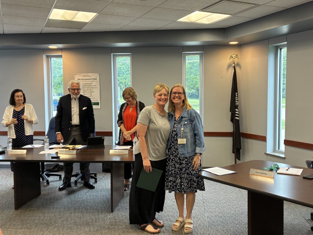 Photograph Broadmoor School Principal wearing a floral navy dress and jean jacket standing next to Broadmoor Adapted Physical Education Teacher as she holds the Ohio Teacher of the Year nomination in a conference room at the Lake County Board of Developmental Disabilities. Both are smiling as people in the background applaud the recognition.