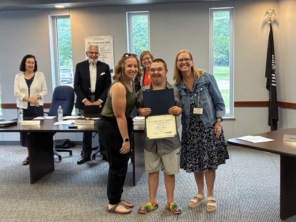 Photograph of a Broadmoor school student wearing a grey shirt and shorts smiling while holding an award certificate for his accomplishments, while two teachers stand on either side of him with three other adults in the background smiling and applauding the recognition in a conference room at the Lake County Board of Developmental Disabilities.