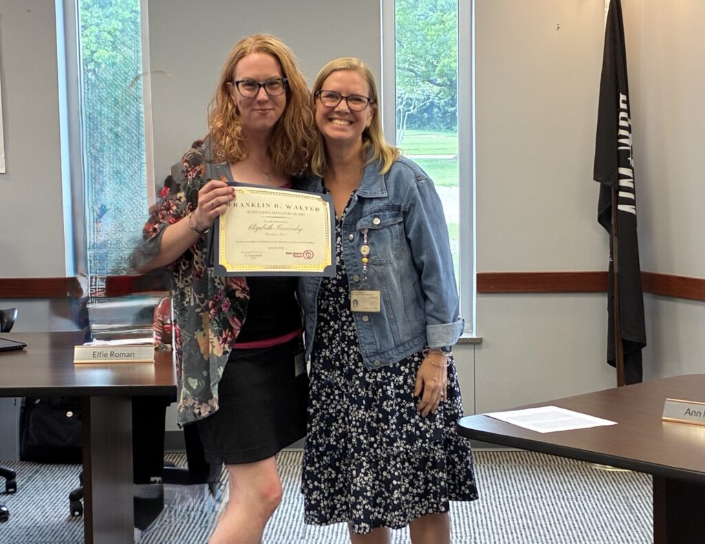 Photograph of Broadmoor School Principal wearing a floral navy dress and jean jacket standing next to Broadmoor Teacher who is wearing a black dress with floral shawl as she holds the "Franklin B Walter Award" in a conference room at the Lake County Board of Developmental Disabilities. Both are smiling and happy.
