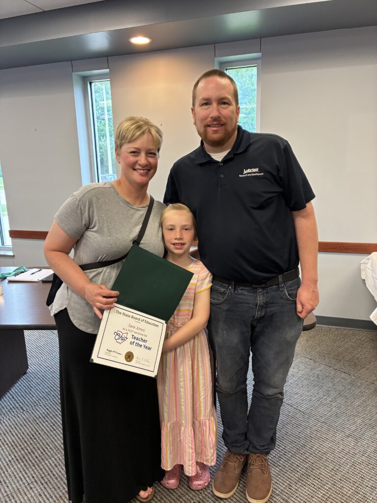 Photograph of a couple standing closely smiling with their young daughter who is wearing a pink striped dress. The woman is a teacher at Broadmoor School and is wearing a grey shirt and black skirt and holding an award saying "Ohio Teacher of the Year". Her husband is wearing a dark blue shirt and blue jeans as the teacher accepts the award in a conference room at the Lake County Board of Developmental Disabilities.