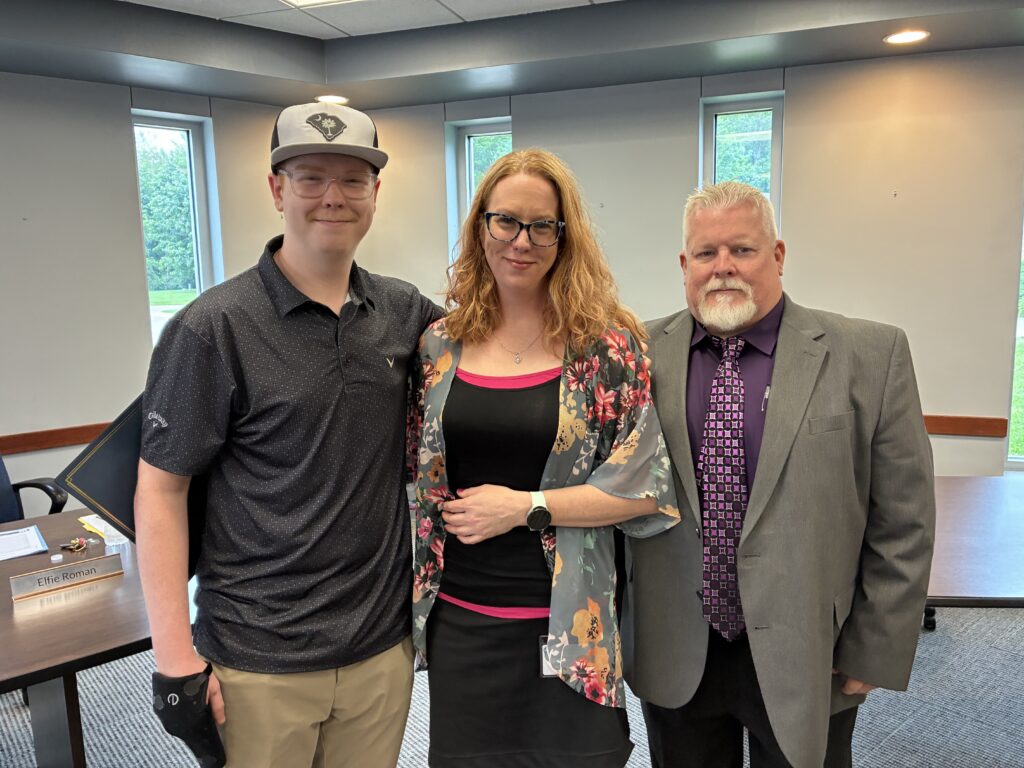 Photograph of Broadmoor Teacher who is wearing a black dress with floral shawl standing closely with her husband and son who are professionally dressed after receiving the "Franklin B Walter Award" in a conference room at the Lake County Board of Developmental Disabilities. All are smiling and happy.