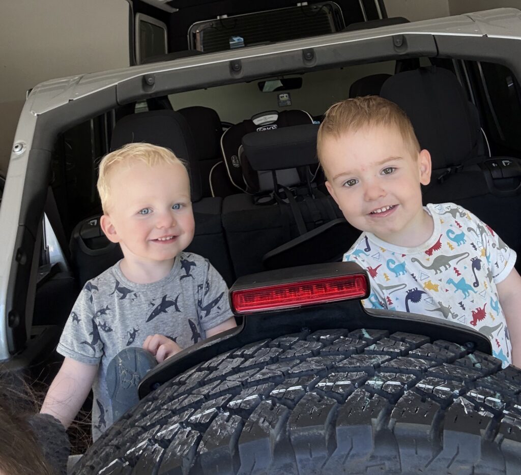 Photo of two toddlers smiling wearing dinosaur and shark t-shirts as they are sitting in the hatch of a car in front of the tire.