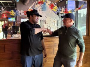 Cory standing beside General Manager Dominic both in restaurant branded shirts and ballcaps doing a fist bump and smiling as they stand inside the Winking Lizard restaurant/pub.
